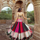 A woman gracefully twirling in a vibrant multi-color printed lehenga choli with intricate mirror work, revealing the stylish back design of the choli with tie-up detailing. The off-white dupatta flows elegantly as she poses in front of a beautifully carved heritage structure, making it a perfect festive outfit for Navratri celebrations.
