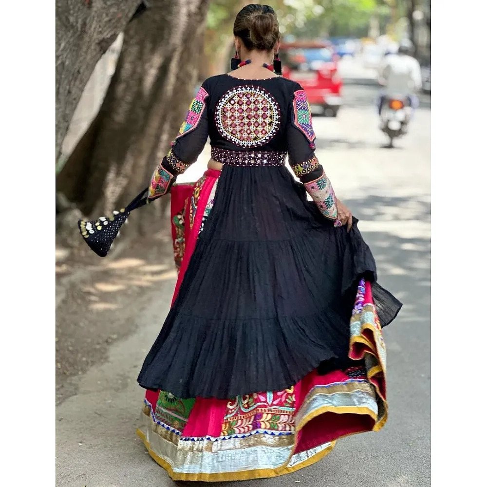 A woman captured from the back, elegantly holding her flowing black and multicolored Gujarati Garba Navratri lehenga chaniya choli, featuring intricate mirror work and embroidery, walking down a street.

