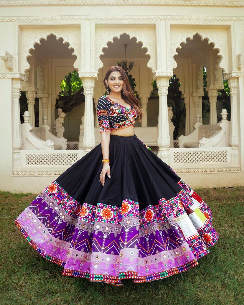 A woman elegantly poses in a black and purple Navratri lehenga choli, featuring intricate Gamthi embroidery and mirror work. She stands in front of a beautifully carved traditional palace backdrop, showcasing the vibrant colors and detailed patterns of her festive outfit.