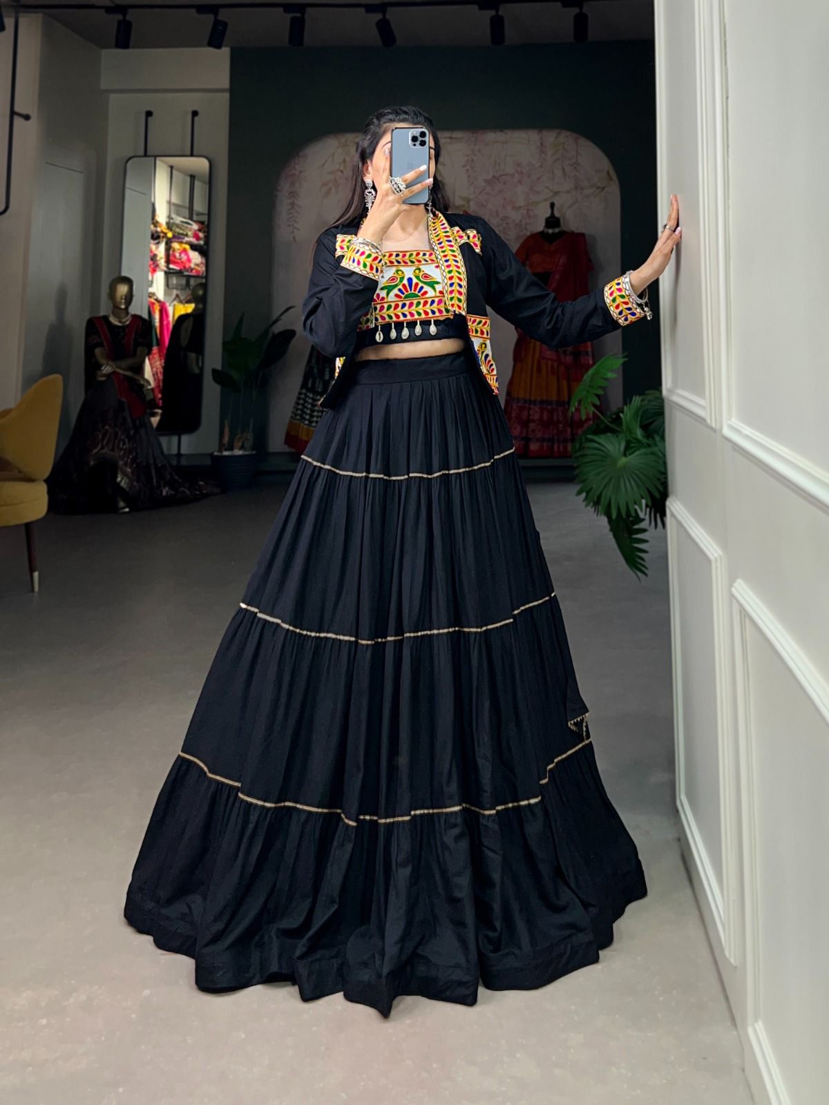 A woman posing in front of a mirror, wearing a black lehenga with gold accents and a colorful embroidered blouse. The boutique setting includes mannequins, dresses, and decorative elements in the background.