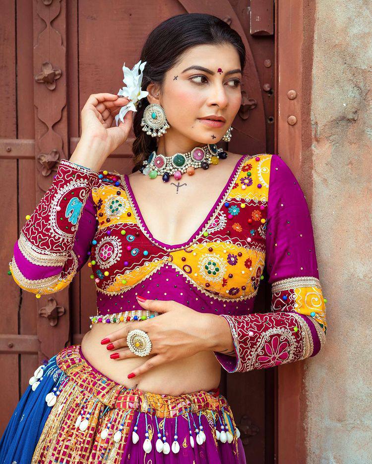 A woman in a vibrant, intricately embroidered Indian lehenga with mirror work and embellishments poses gracefully against a rustic door, adorned with statement jewelry and a floral hair accessory.
