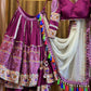 A stunning magenta lehenga set hanging on wooden hangers, featuring intricate mirror work, embroidery, and vibrant tassel details. The outfit includes a flared skirt with colorful motifs, a matching blouse with detailed sleeves, and a sheer white dupatta with pearl embellishments and multicolored tassels. The rich ethnic design and craftsmanship highlight the beauty of traditional Indian attire.