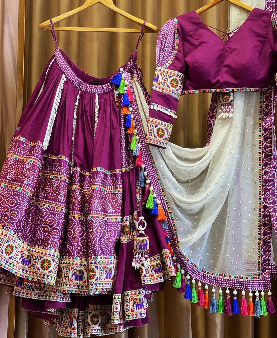 A stunning magenta lehenga set hanging on wooden hangers, featuring intricate mirror work, embroidery, and vibrant tassel details. The outfit includes a flared skirt with colorful motifs, a matching blouse with detailed sleeves, and a sheer white dupatta with pearl embellishments and multicolored tassels. The rich ethnic design and craftsmanship highlight the beauty of traditional Indian attire.