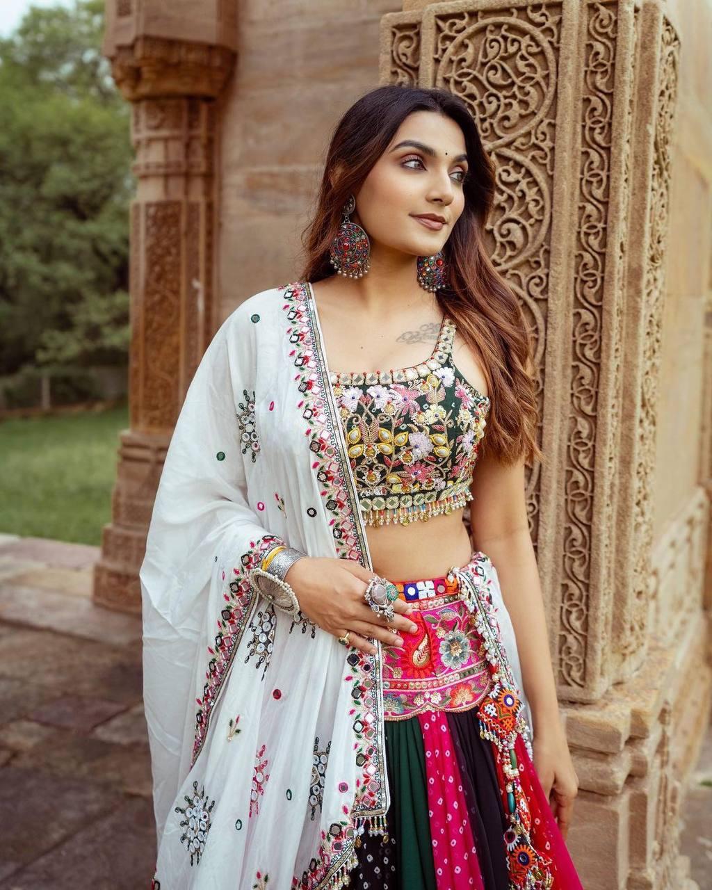 A woman poses gracefully in a vibrant multicolor lehenga choli, featuring intricate mirror work and floral embroidery. She pairs the outfit with a beautifully embellished white dupatta and statement jewelry, including large earrings and rings. Standing against a traditional carved sandstone structure, she exudes elegance and festive charm, making this a perfect look for Navratri or Indian cultural celebrations.