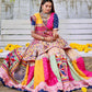 A woman sits gracefully on a bench wearing a colorful Navratri lehenga with intricate digital prints, mirror work, and embroidery, surrounded by marigold flowers.