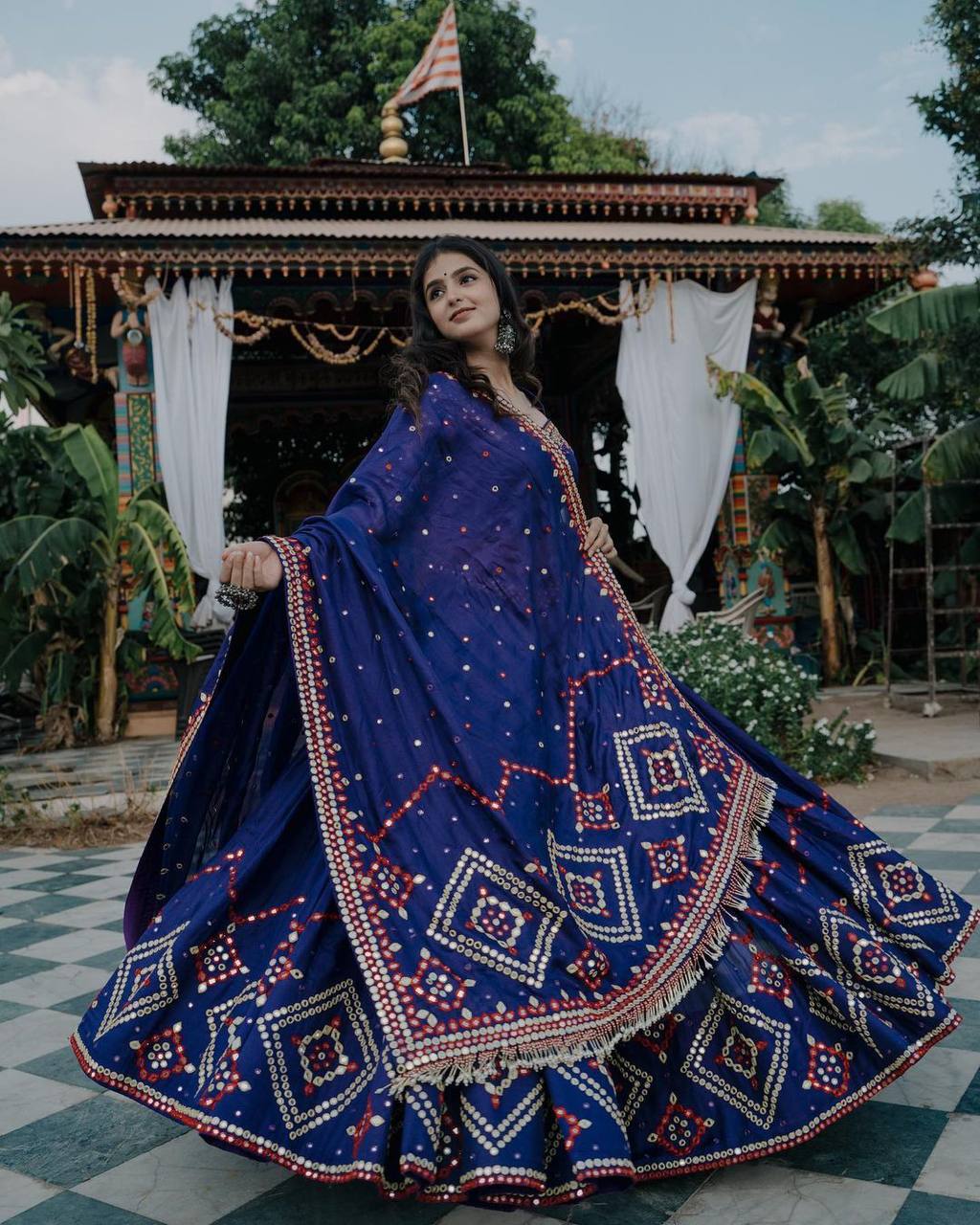 A woman gracefully twirling in a royal blue lehenga adorned with intricate embroidery and mirror work, set against a beautifully decorated traditional backdrop.