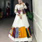 A woman poses in a stunning white lehenga with multicolored patchwork panels, mirror work, and intricate embroidery. She pairs it with a fitted blouse and a decorative mirror-work jacket, accessorized with statement silver jewelry, standing barefoot in a boutique setting.