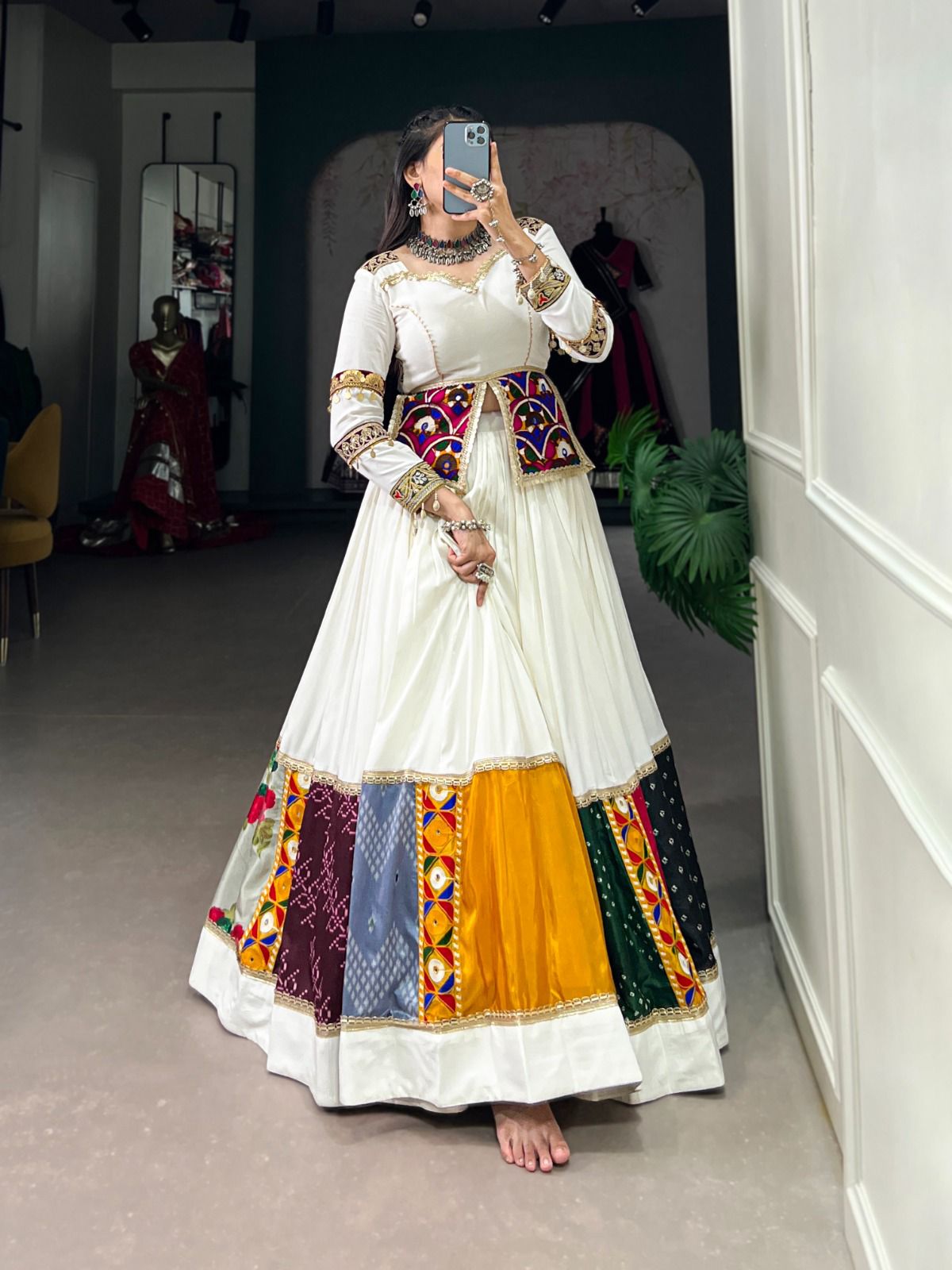 A woman poses in a stunning white lehenga with multicolored patchwork panels, mirror work, and intricate embroidery. She pairs it with a fitted blouse and a decorative mirror-work jacket, accessorized with statement silver jewelry, standing barefoot in a boutique setting.