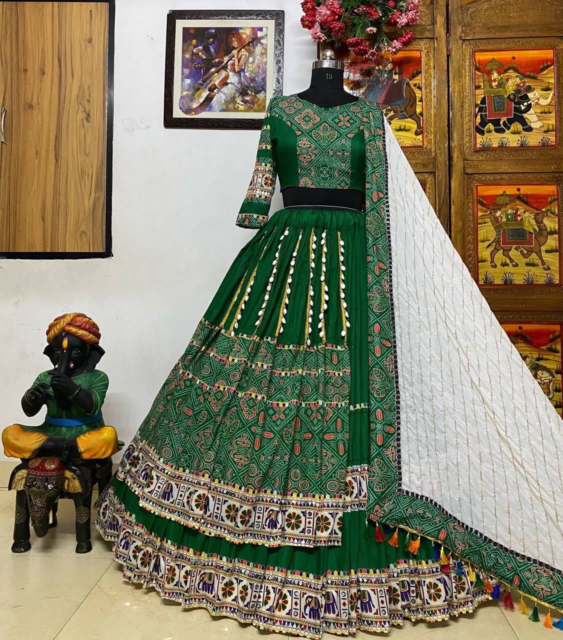 A stunning green Gujarati lehenga displayed on a mannequin, featuring intricate mirror work, geometric embroidery, and a heavily embroidered border with folk and elephant motifs. The lehenga is paired with a matching blouse with three-quarter sleeves and a white dupatta adorned with colorful tassels and embroidered detailing. The outfit is set against a traditional backdrop, including a decorative statue of Lord Krishna and Rajasthani-style artwork, adding to its cultural elegance.