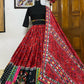  A beautifully designed traditional red embroidered lehenga choli with mirror work and multicolored patchwork is displayed on a mannequin. The outfit features a black blouse with elbow-length sleeves and a matching dupatta draped elegantly over the lehenga, set against a wooden door and decorative interior.