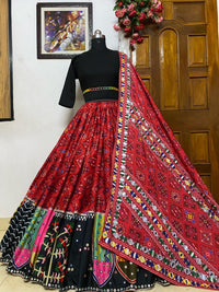  A beautifully designed traditional red embroidered lehenga choli with mirror work and multicolored patchwork is displayed on a mannequin. The outfit features a black blouse with elbow-length sleeves and a matching dupatta draped elegantly over the lehenga, set against a wooden door and decorative interior.