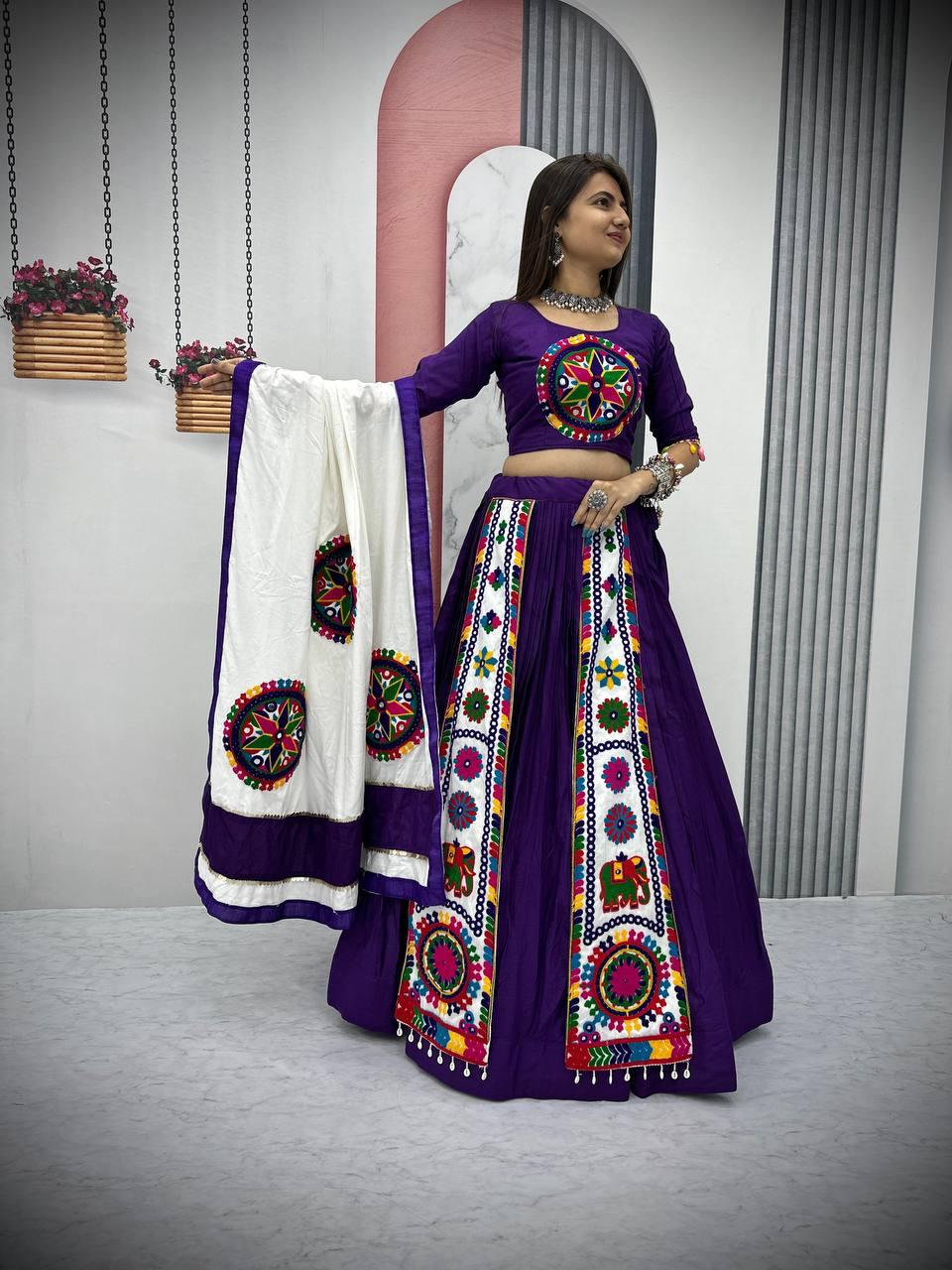A woman elegantly poses in a stunning purple Navratri lehenga choli featuring intricate Gamthi embroidery and mirror work. The lehenga showcases vibrant traditional motifs, including elephants and geometric patterns, complemented by a matching blouse with detailed embroidery. She gracefully holds a contrasting white dupatta adorned with circular embroidered designs and a deep purple border, enhancing the festive appeal. Styled with traditional silver jewelry and bangles, the outfit is perfect for Garba and 