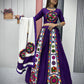 A woman gracefully showcases a vibrant purple Navratri lehenga choli adorned with intricate Gamthi embroidery. The outfit features colorful traditional motifs, mirror work, and a matching blouse with detailed patterns. She holds a contrasting white dupatta with embroidered circular designs, complementing the festive look. The elegant jewelry and bangles enhance the ethnic charm, making it a perfect attire for Garba and Navratri celebrations.