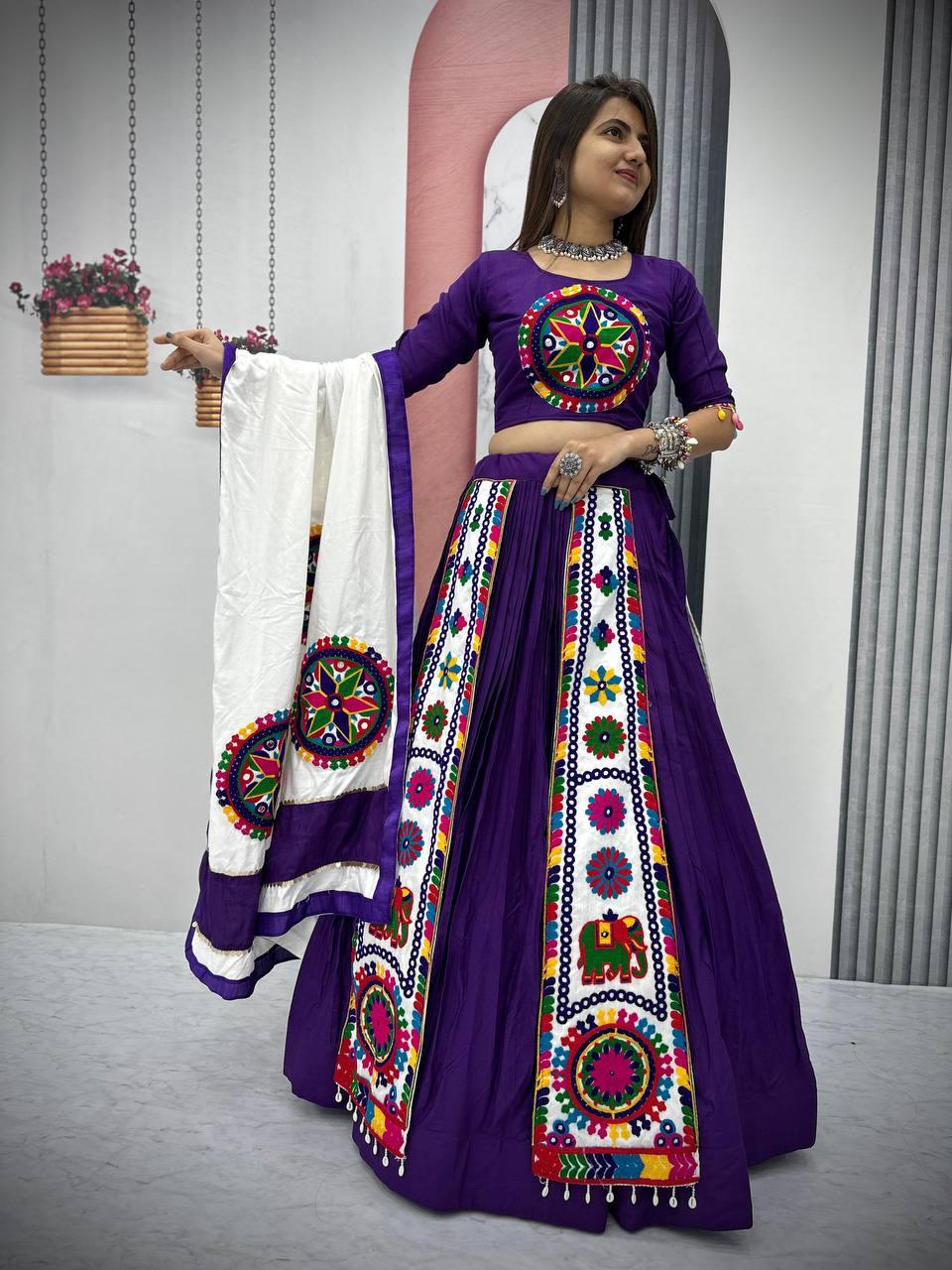 A woman gracefully showcases a vibrant purple Navratri lehenga choli adorned with intricate Gamthi embroidery. The outfit features colorful traditional motifs, mirror work, and a matching blouse with detailed patterns. She holds a contrasting white dupatta with embroidered circular designs, complementing the festive look. The elegant jewelry and bangles enhance the ethnic charm, making it a perfect attire for Garba and Navratri celebrations.