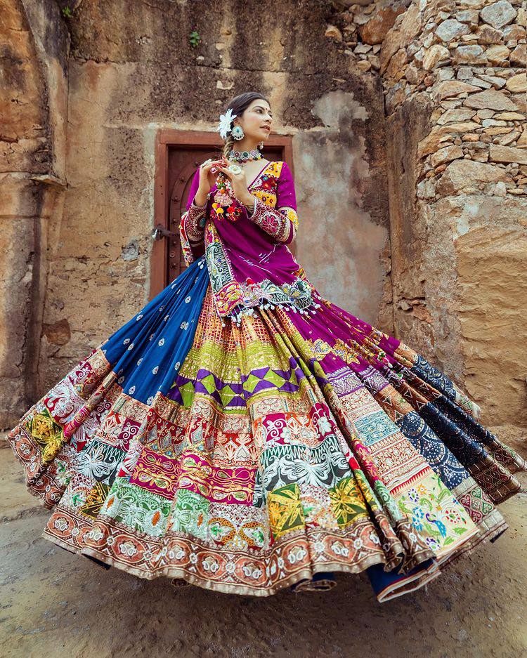 A woman elegantly twirls in a multicolored, intricately designed Indian lehenga with detailed embroidery and mirror work, set against the backdrop of an ancient rustic structure.