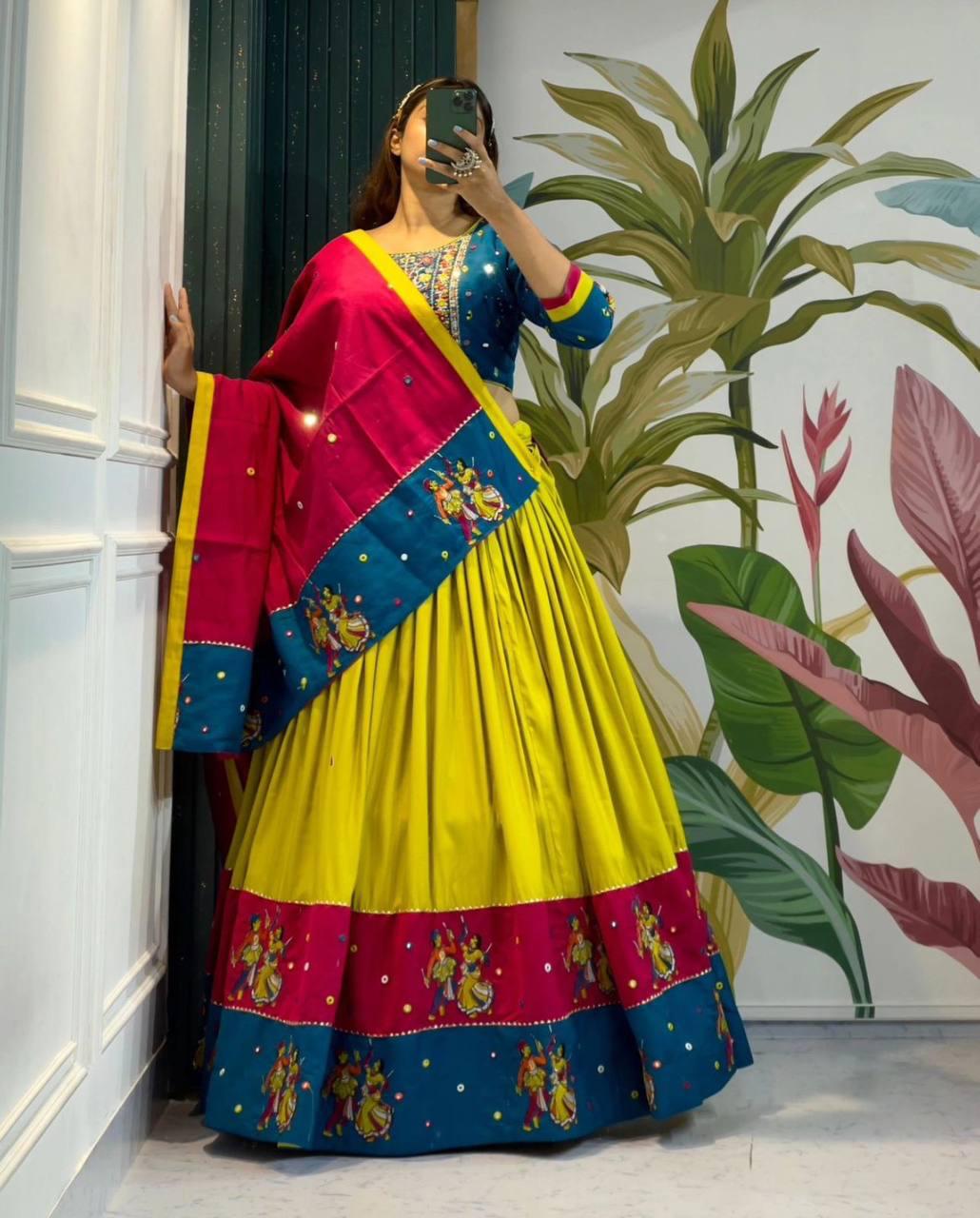 A woman wearing a vibrant traditional Gujarati lehenga choli set, featuring a bright yellow skirt with a blue and pink embroidered border, a blue blouse with mirror work, and a matching dupatta. She is taking a mirror selfie in a stylish room with botanical wall art, showcasing the festive and cultural essence of Navratri attire.