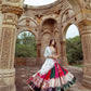 A woman wearing a vibrant multi-color printed lehenga choli with an off-white dupatta, adorned with real mirror work and intricate embroidery, posing elegantly in front of an ancient heritage structure with carved arches. Perfect festive wear for Navratri celebrations.