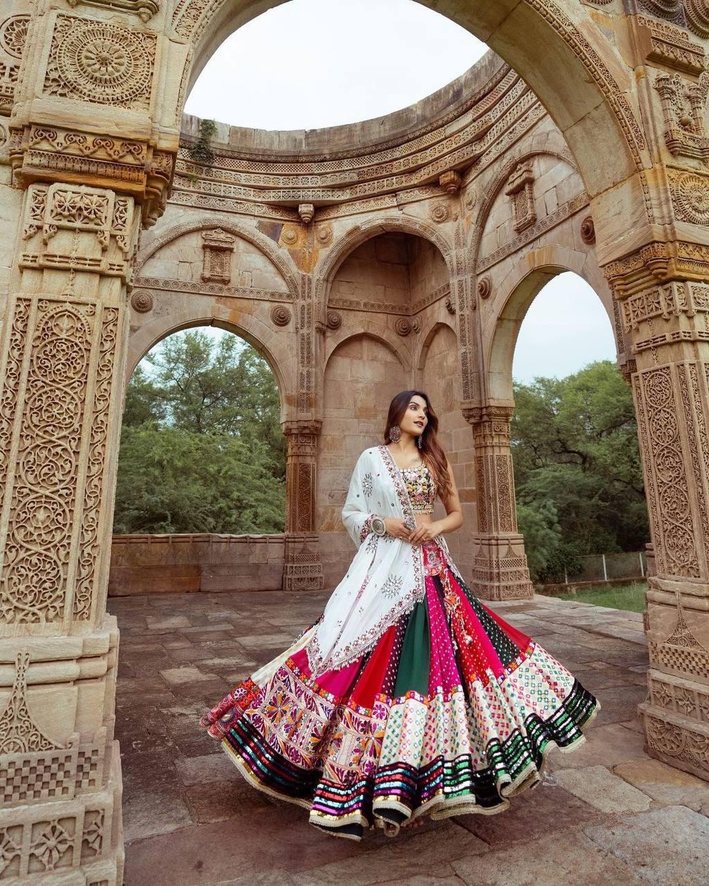 A woman wearing a vibrant multi-color printed lehenga choli with an off-white dupatta, adorned with real mirror work and intricate embroidery, posing elegantly in front of an ancient heritage structure with carved arches. Perfect festive wear for Navratri celebrations.