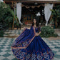 A woman wearing a stunning navy blue lehenga choli with intricate embroidery and mirror work, posing elegantly in a traditional setting with rich décor.