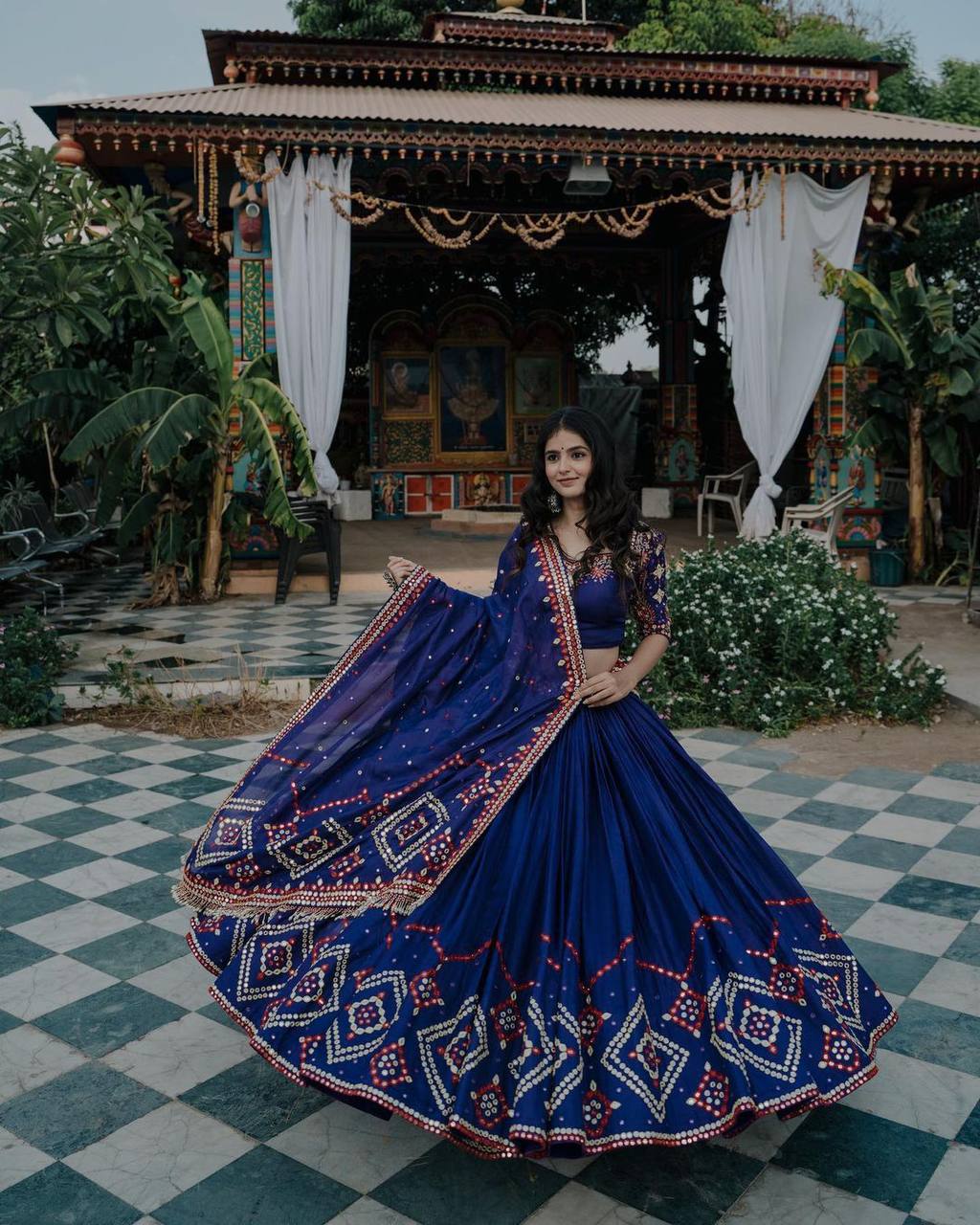 A woman wearing a stunning navy blue lehenga choli with intricate embroidery and mirror work, posing elegantly in a traditional setting with rich décor.