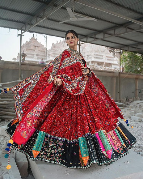 A woman elegantly twirls in a red traditional Navratri lehenga choli adorned with intricate mirror work, vibrant embroidery, and a flared silhouette, set against a rustic backdrop with a temple in the distance.