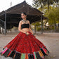 A woman poses gracefully in a vibrant red embroidered Navratri lehenga choli with mirror work and a flared silhouette, paired with a stylish black blouse, set against a rustic outdoor backdrop.