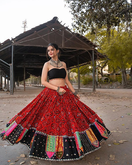 A woman poses gracefully in a vibrant red embroidered Navratri lehenga choli with mirror work and a flared silhouette, paired with a stylish black blouse, set against a rustic outdoor backdrop.