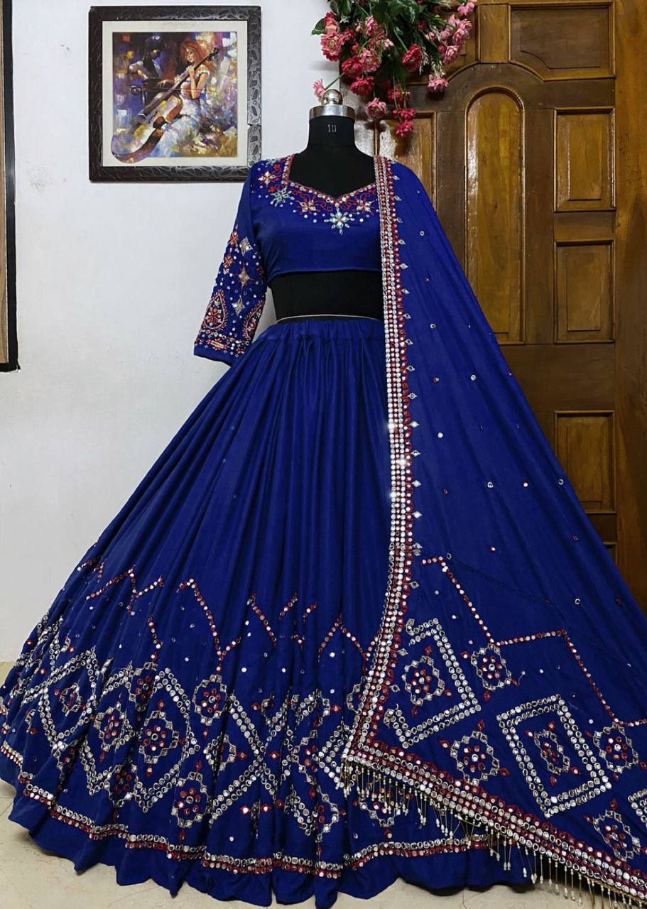 A beautifully designed royal blue lehenga with intricate embroidery and mirror work, elegantly displayed on a mannequin against a wooden door and a framed artwork in the background.