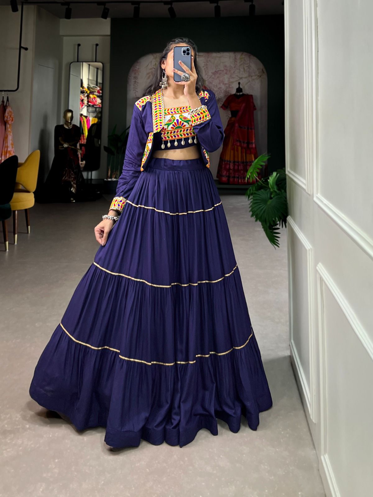 A woman poses in front of a mirror wearing a navy blue lehenga with gold detailing, paired with a colorful embroidered blouse and matching jacket. The boutique setting features mannequins, designer outfits, and elegant decor in the background.