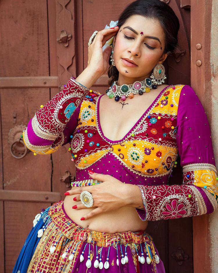 A woman in a vibrant, intricately embroidered Indian lehenga poses gracefully against a rustic wooden door, adorned with statement jewelry and delicate makeup, exuding regal charm.