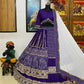 A stunning purple Bandhani lehenga displayed on a mannequin, featuring intricate white tie-dye patterns and colorful embroidered borders with elephant motifs. The lehenga is paired with a matching blouse adorned with decorative mirror work and tassels, and a contrasting white dupatta with delicate embroidery and multicolored tassels. The setup is complemented by traditional Indian décor, including paintings and a statue, enhancing the ethnic charm.