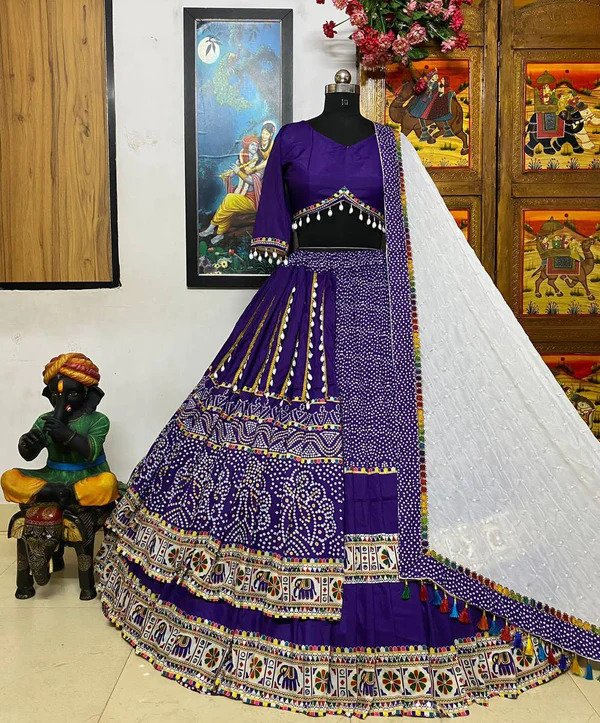 A stunning purple Bandhani lehenga displayed on a mannequin, featuring intricate white tie-dye patterns and colorful embroidered borders with elephant motifs. The lehenga is paired with a matching blouse adorned with decorative mirror work and tassels, and a contrasting white dupatta with delicate embroidery and multicolored tassels. The setup is complemented by traditional Indian décor, including paintings and a statue, enhancing the ethnic charm.
