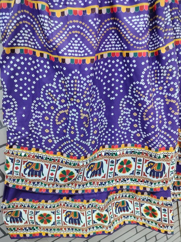 A vibrant purple fabric featuring intricate white Bandhani (tie-dye) patterns in symmetrical designs. The fabric is adorned with colorful decorative borders, including multicolored tassel-like embroidery and a detailed elephant motif strip with floral elements. The traditional design and rich colors make it ideal for ethnic wear.