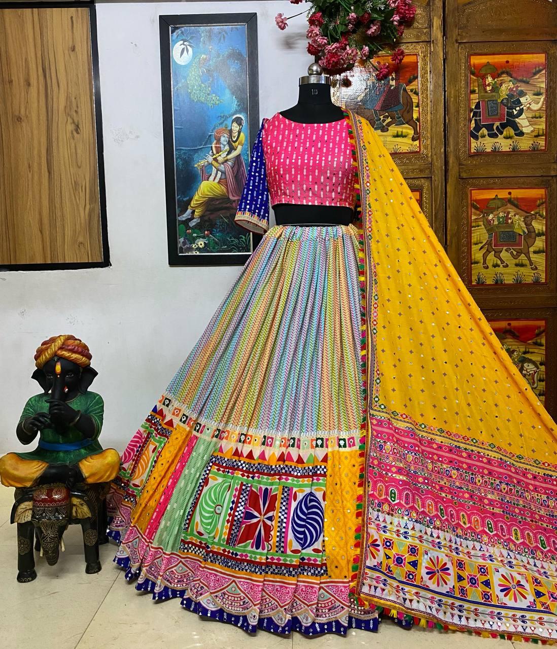 A stunning Gujarati lehenga displayed on a mannequin, featuring a multicolored skirt with intricate patchwork and embroidery, a pink and blue blouse with mirror work, and a bright yellow dupatta adorned with traditional embellishments. The setup includes a Radha-Krishna painting and a decorative statue, enhancing the cultural aesthetic.