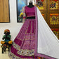 A beautifully designed magenta lehenga displayed on a mannequin, featuring intricate mirror work, embroidery, and traditional motifs. The outfit includes a matching blouse with embellished sleeves and a contrasting white dupatta adorned with delicate pearls and colorful tassels. The backdrop showcases vibrant Rajasthani decor with wooden carvings and paintings, enhancing the cultural aesthetic.
