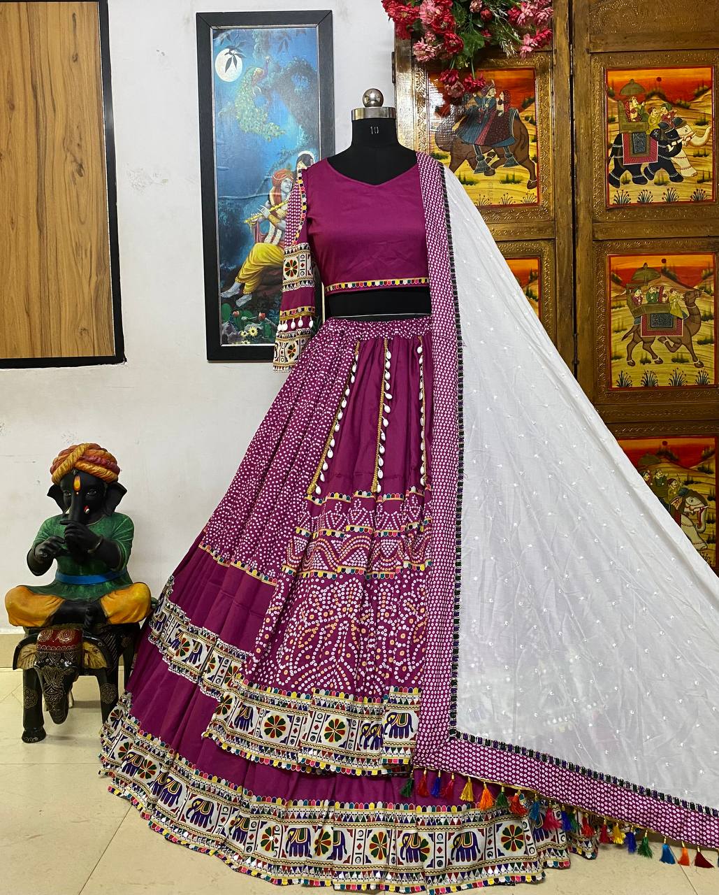 A beautifully designed magenta lehenga displayed on a mannequin, featuring intricate mirror work, embroidery, and traditional motifs. The outfit includes a matching blouse with embellished sleeves and a contrasting white dupatta adorned with delicate pearls and colorful tassels. The backdrop showcases vibrant Rajasthani decor with wooden carvings and paintings, enhancing the cultural aesthetic.
