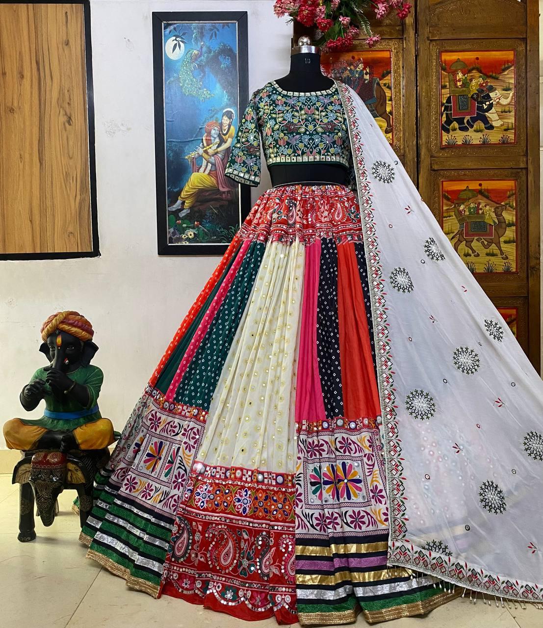 A beautifully designed multicolor lehenga choli displayed on a mannequin, featuring intricate embroidery, mirror work, and a vibrant patchwork design. The outfit is paired with a heavily embellished blouse and a white dupatta adorned with delicate embroidery. The background includes traditional Indian décor, adding to the ethnic charm, making this an ideal outfit for Navratri, Garba, or festive celebrations.