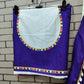 A neatly folded deep purple blouse piece with a white circular design in the center, bordered by vibrant multicolored tassel-like embroidery. The bottom edge features a similar colorful border. The fabric is displayed on a hanger against a brick-patterned background, showcasing its ethnic and festive appeal.