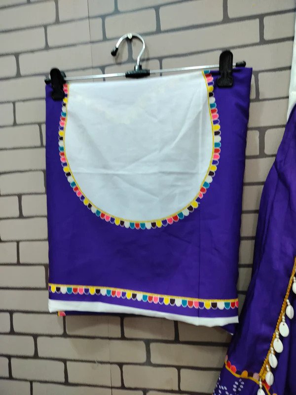 A neatly folded deep purple blouse piece with a white circular design in the center, bordered by vibrant multicolored tassel-like embroidery. The bottom edge features a similar colorful border. The fabric is displayed on a hanger against a brick-patterned background, showcasing its ethnic and festive appeal.