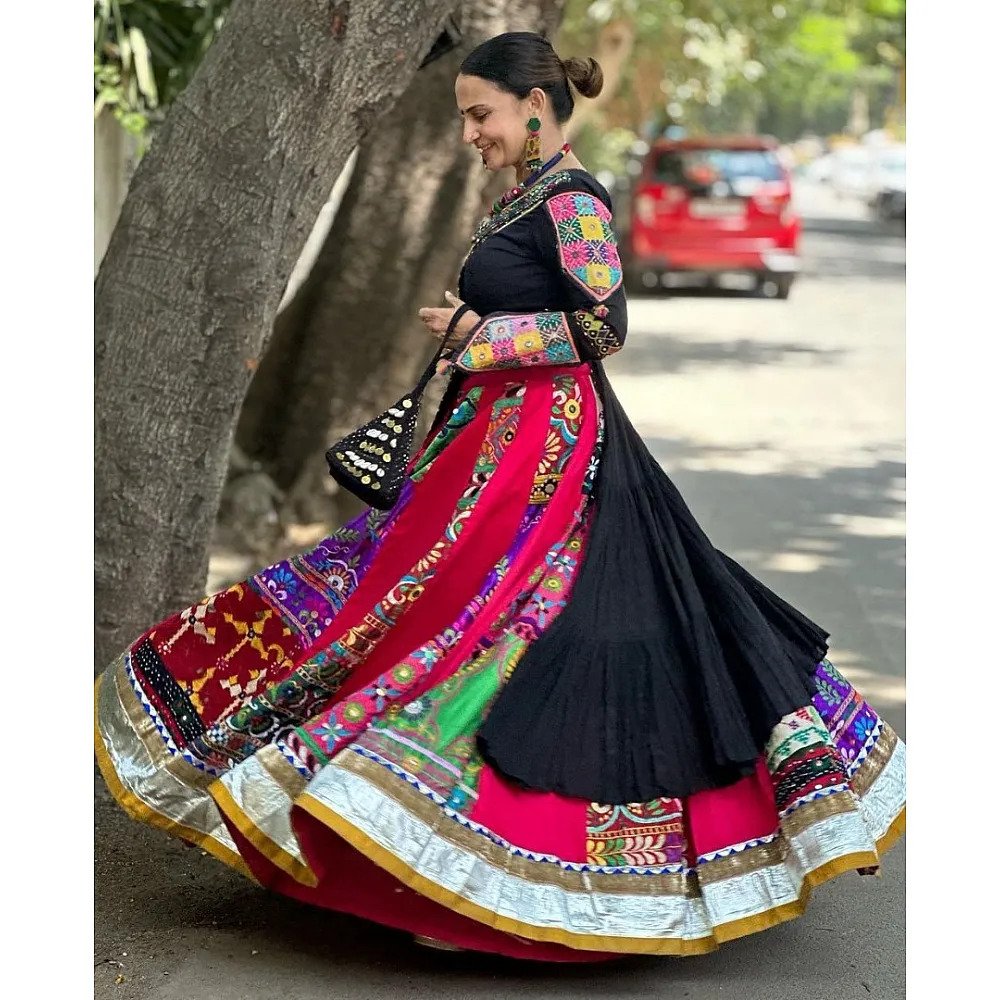 A woman gracefully twirling in a vibrant black and multicolored Gujarati Garba Navratri lehenga chaniya choli with intricate mirror work and embroidery, outdoors with a joyful expression.

