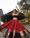  A woman elegantly twirls in a traditional red embroidered Navratri lehenga choli adorned with mirror work and multicolored patchwork, paired with a black blouse. She poses gracefully with raised arms, showcasing her ethnic jewelry, against a rustic outdoor setting.