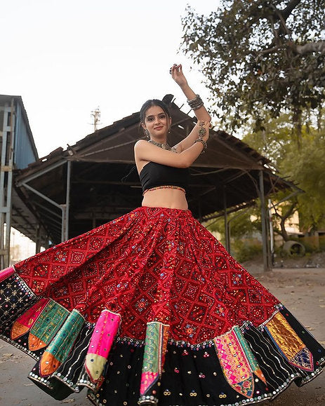  A woman elegantly twirls in a traditional red embroidered Navratri lehenga choli adorned with mirror work and multicolored patchwork, paired with a black blouse. She poses gracefully with raised arms, showcasing her ethnic jewelry, against a rustic outdoor setting.