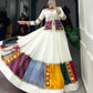 A woman elegantly twirls in a white lehenga with multicolored patchwork panels, mirror work, and intricate embroidery. She is wearing a matching blouse and a vibrant mirror-work jacket, accessorized with traditional silver jewelry, in a boutique setting.