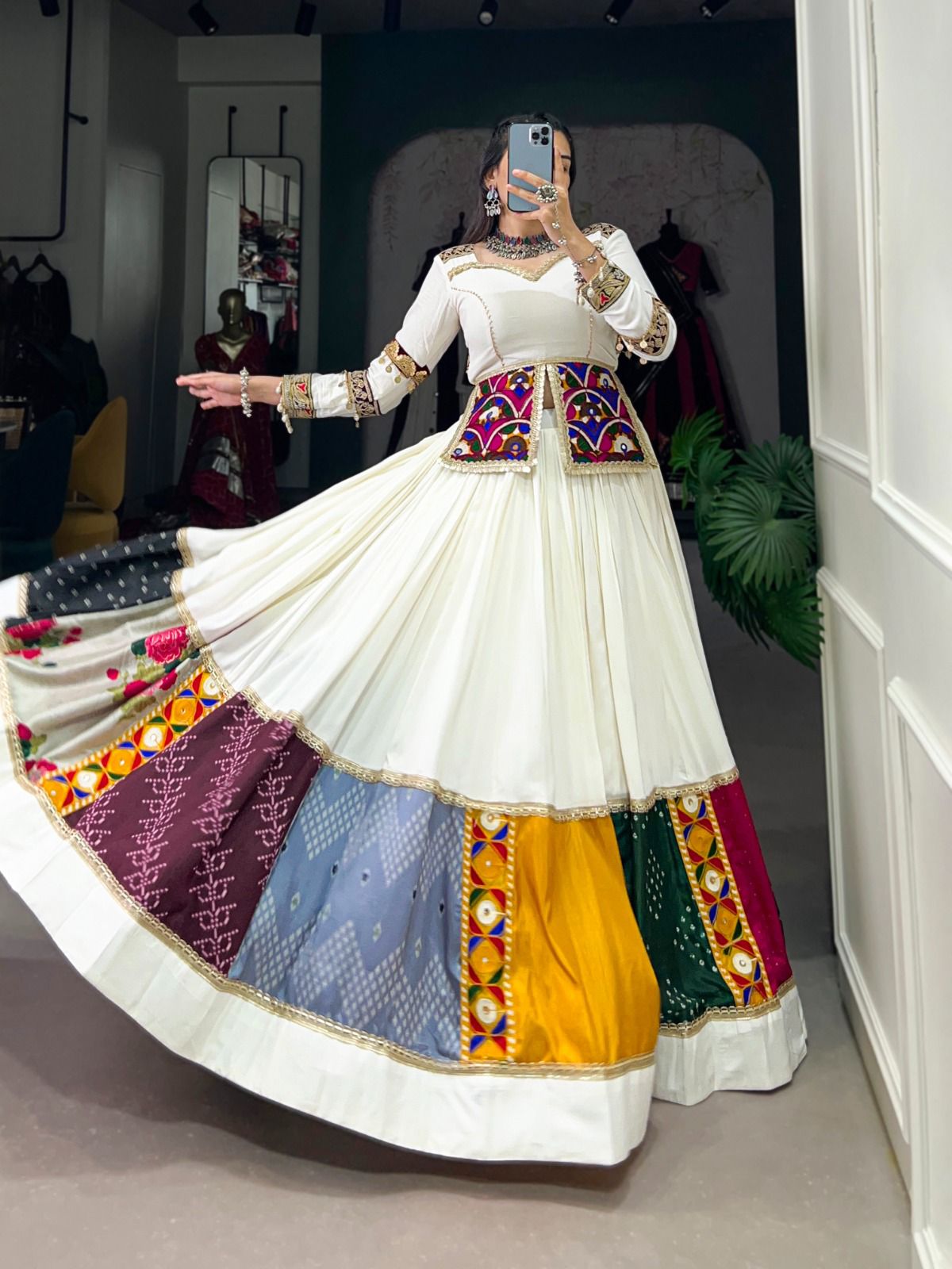 A woman elegantly twirls in a white lehenga with multicolored patchwork panels, mirror work, and intricate embroidery. She is wearing a matching blouse and a vibrant mirror-work jacket, accessorized with traditional silver jewelry, in a boutique setting.