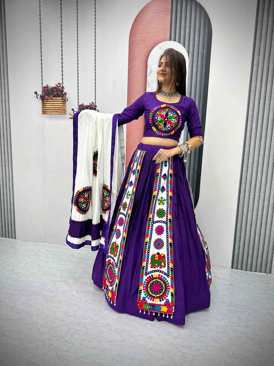 Woman wearing a purple Navratri lehenga with colorful Gamthi embroidery, holding a white dupatta with matching mirror work.
