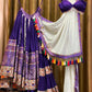 A gorgeous deep purple lehenga set hanging on wooden hangers, adorned with traditional embroidery, mirror work, and multicolored tassels. The outfit features a flared skirt with detailed motifs, a stylish blouse with thin straps, and a sheer white dupatta with colorful tassel embellishments. The ensemble showcases exquisite craftsmanship and rich ethnic aesthetics, perfect for festive and cultural celebrations.