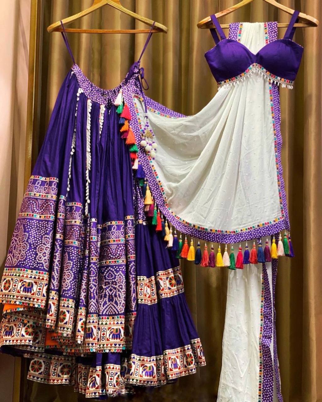 A gorgeous deep purple lehenga set hanging on wooden hangers, adorned with traditional embroidery, mirror work, and multicolored tassels. The outfit features a flared skirt with detailed motifs, a stylish blouse with thin straps, and a sheer white dupatta with colorful tassel embellishments. The ensemble showcases exquisite craftsmanship and rich ethnic aesthetics, perfect for festive and cultural celebrations.