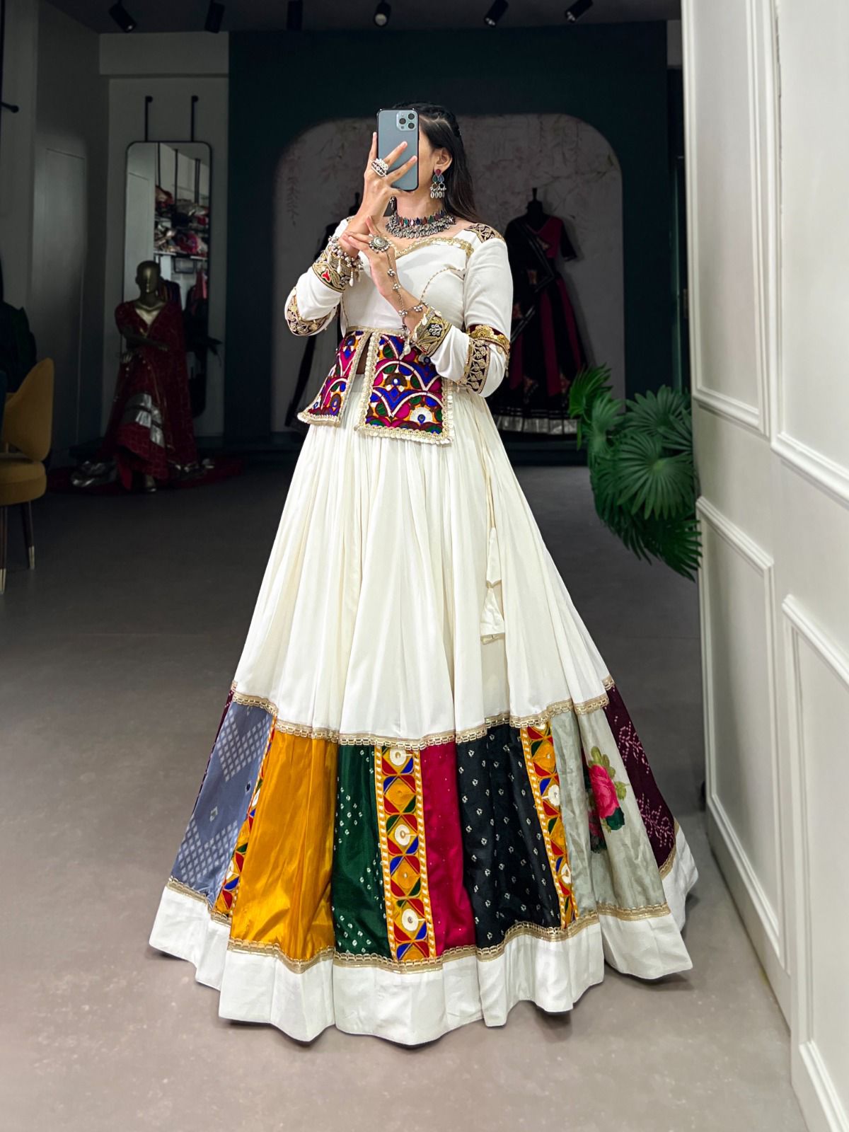 A woman wearing a white lehenga adorned with multicolored patchwork panels at the hem, featuring intricate mirror work and embroidery. She pairs it with a matching blouse and a vibrant mirror-work jacket, accessorized with silver traditional jewelry, while capturing her look in a boutique setting.







