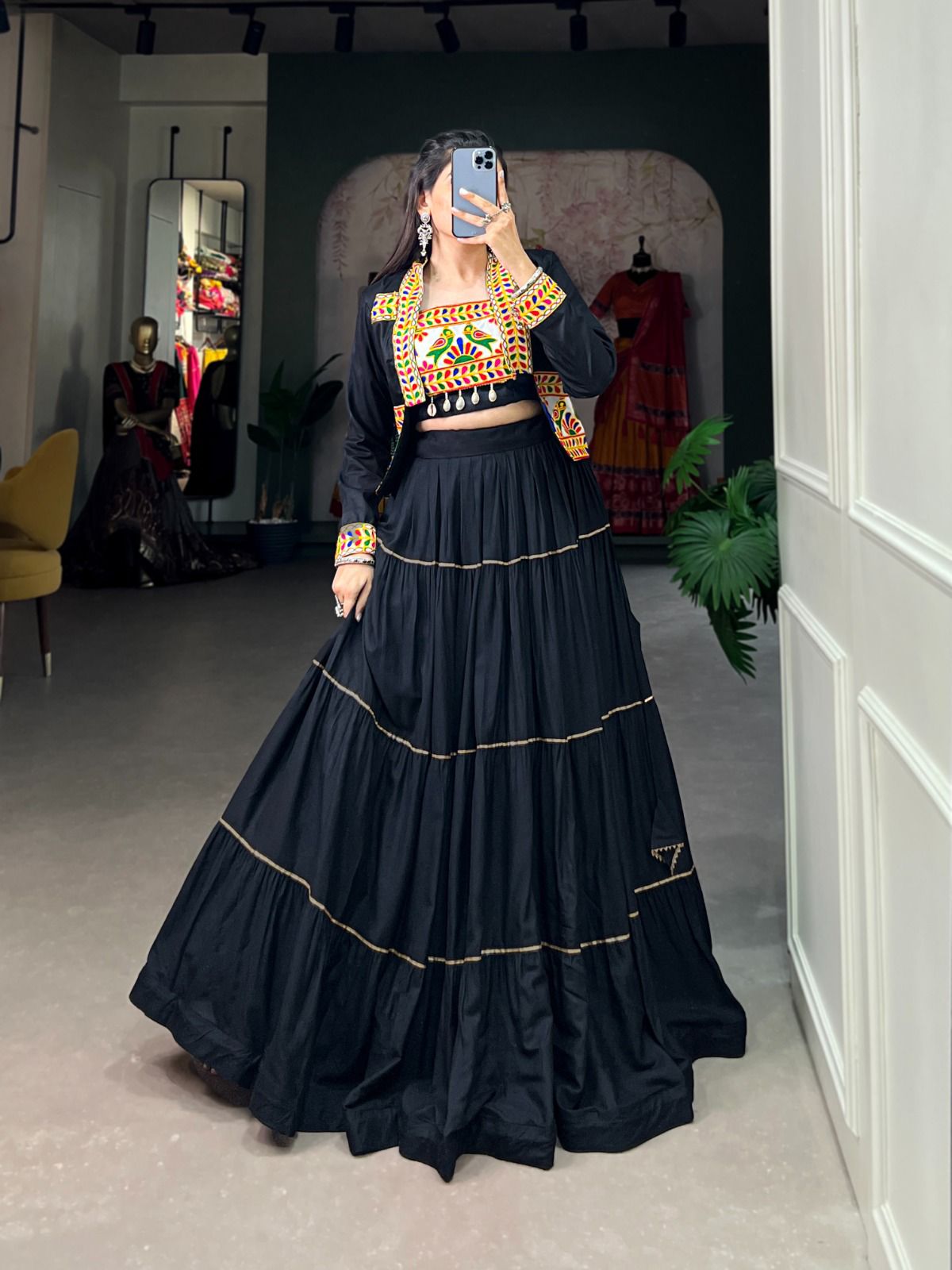 A woman taking a mirror selfie in a boutique, wearing a black lehenga with gold detailing and a vibrant, multicolored embroidered blouse. The background features a stylish interior with mannequins, dresses, and decor elements.