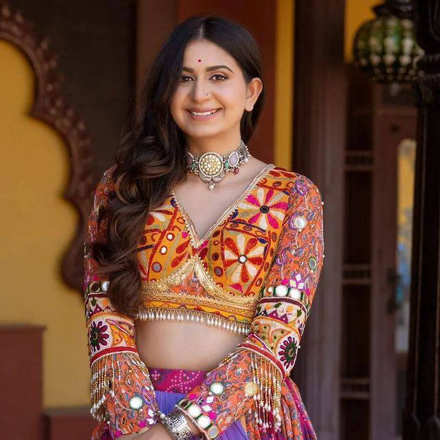 A smiling woman wearing a traditional Gujarati lehenga choli with intricate embroidery, mirror work, and colorful patterns. She is accessorized with a statement choker necklace and bangles, standing in a heritage-style setting.
